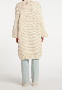 Dikke gebreide crèmekleurige cardigan met een losse pasvorm, voorzien van geribbelde manchetten en zoom. Gecombineerd met lichtblauwe jeans en witte schoenen.