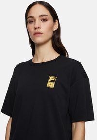 Zwarte oversized t-shirt gemaakt van katoen, met een goudkleurig logo patch op de linkerborst en korte mouwen.