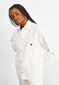 Veste en toile blanche avec des boutons-pression en argent, deux poches poitrine et un patch logo. Présente une coupe décontractée et une texture lisse.