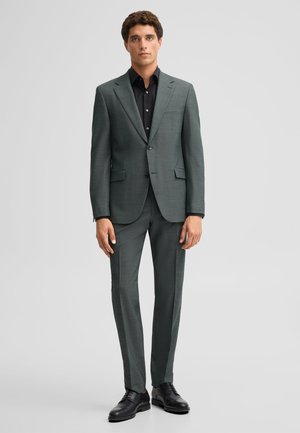 Man stands facing forward wearing a dark gray tailored suit with a black dress shirt and black dress shoes on a plain background.