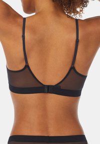 Schiena di una donna che indossa un reggiseno trasparente nero con spalline regolabili e chiusura a gancio, su uno sfondo chiaro e uniforme.