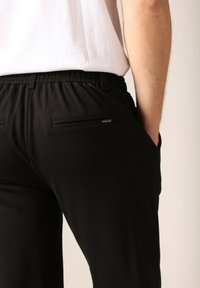 Zwarte broek met een elastische tailleband, voorzien van twee achterzakken met horizontale openingen. Gemaakt van een soepele stof met een strakke uitstraling.