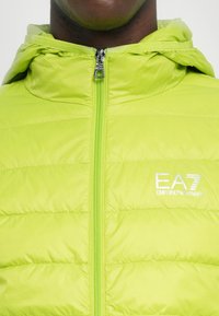 Jakna od pufera u limeta zelenoj boji s kapuljačom, s prepoznatljivim kvadrom, prednjim patentnim zatvaračem i EA7 Emporio Armani logotipom na prsima.