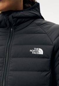 Giacca a vento nera con colletto alto e cuciture visibili. Presenta un logo bianco sul petto e una zip frontale per la chiusura.