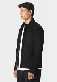 Černá nubuková bunda s límcem, zapínáním na knoflíky vpředu a dvěma bočními kapsami. Nositelná přes bílou košili, vystavená na jednolitém pozadí.