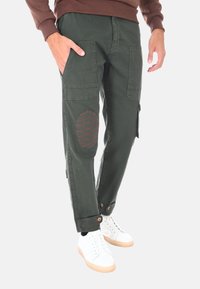 Pantaloni cargo verde scuro con ampie tasche frontali, toppe sulle ginocchia con cuciture rosse, orli regolabili e vestibilità rilassata. Abbinati a sneakers bianche.