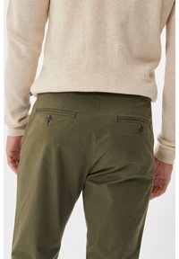 Olive green pants with a smooth texture, featuring two back pockets with button closures, worn with a beige knit sweater.
