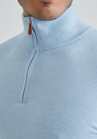 Pull à col montant bleu clair avec demi-fermeture éclair. Texture en tricot doux, avec une tirette en cuir contrastée sur la fermeture éclair.
