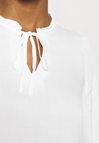 Blusa bianca realizzata in tessuto leggero, caratterizzata da un collo arricciato e un'apertura a occhiello con dettaglio in legatura nella parte anteriore.