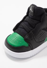 Černá a zelená dětská teniska s bílou podrážkou, perforovanou špičkou a bočními panely, černými elastickými tkaničkami a logem Jumpman na jazyku.