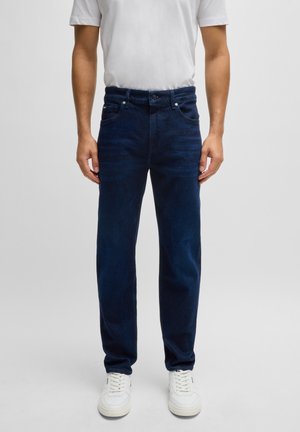 Mężczyzna ubrany w ciemnoniebieskie jeansy o prostym kroju, białe trampki i białą koszulkę z krótkim rękawem, stojący na tle jednolitego szarego tła.