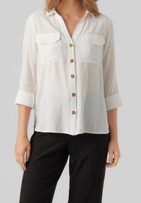 Camicia bianca con bottoni, dotata di due tasche sul petto, con colletto e maniche arrotolate. Tessuto liscio con una texture semi-trasparente.