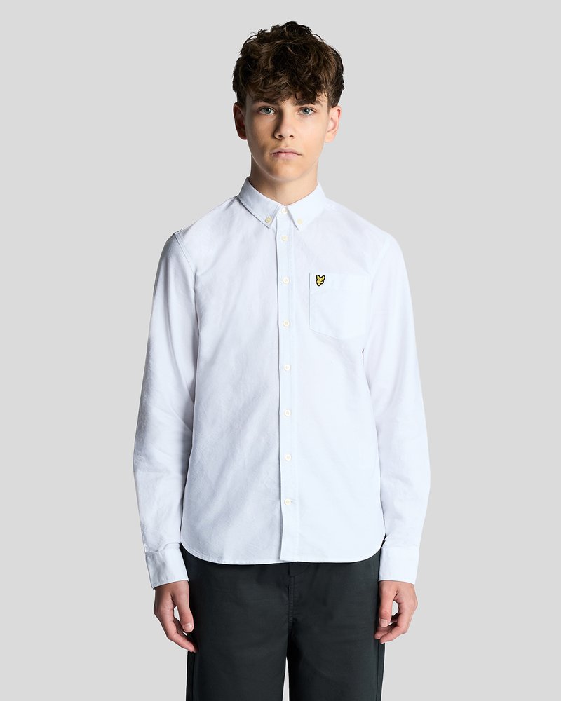 Νέος άνδρας με σγουρά μαλλιά, που φοράει ένα λευκό μακρυμάνικο πουκάμισο με κουμπιά και ένα μικρό κίτρινο λογότυπο στην τσέπη του στήθους, στέκεται μπροστά σε γκρι φόντο.