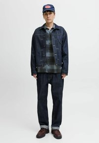 Chaqueta de mezclilla oscura con costuras amarillas, superpuesta sobre una camisa de cuadros verde y azul. Jeans oscuros holgados y zapatos marrones con una gorra azul.