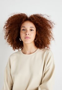Beige Sweatshirt mit übergroßem Schnitt und Rundhalsausschnitt, aus weichem Material. Das Model hat lockiges rotes Haar und Ohrringe in Form von Ringen. Heller Hintergrund.