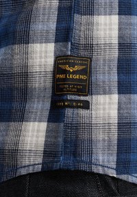 Blauw en wit geruit stof met een genaaide label met "PME LEGEND," gouden letters en een ontwerp met een vleugelsymbool.