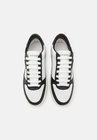 Sneaker bianco e nero con tomaia in pelle liscia, punta rotonda, dettagli perforati e lacci bianchi piatti. Nome del marchio "GUESS" stampato sulla linguetta.