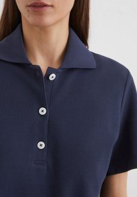 Navyblaues Poloshirt aus strukturiertem Stoff mit gewickeltem Kragen, vier weißen Knöpfen und einer kleinen Knopfleiste. Kurzarm vervollständigt das Design.