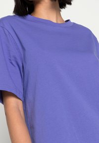 Fioletowa bawełniana koszulka z krótkim rękawem i okrągłym dekoltem; gładka tekstura i minimalne detale szycia, widok pod kątem ukazujący układ materiału.