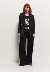 Blazer nero a righe con bottoni argentati, indossato su una t-shirt nera con grafica floreale. Abbinato a pantaloni neri a gamba larga e scarpe con stampa leopardata.