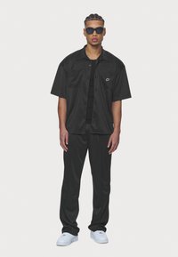 Black short-sleeve button-up shirt with two pockets, paired with black pants. Worn with sunglasses and white sneakers. Smooth texture.