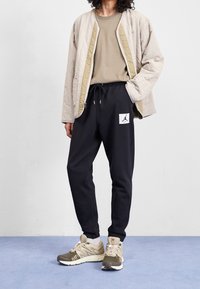 Beige vadderad jacka, beige skjorta, svarta joggingbyxor med vit logotyp och beige-gröna sneakers. Avslappnad sportoutfit med fickdetaljer.