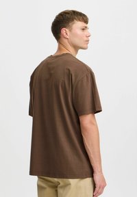 Camiseta de algodón marrón con un ajuste holgado, mangas cortas y cuello redondo. La tela parece suave con costuras mínimas.