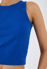 Geribbelde, mouwloze, cropped top in een levendig blauw. Het heeft een ronde halslijn en een zachte textuur, wat de rek en pasvorm van de stof benadrukt.