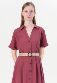 Robe chemise bordeaux à manches courtes retroussées, taille cintrée et ceinture tressée de couleur claire avec une boucle circulaire.