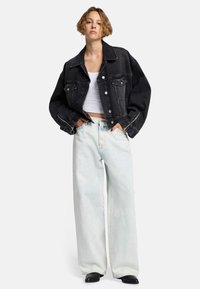 Schwarze Denim-ähnliche Oversized-Jacke mit Fronttaschen, kombiniert mit hellblauen weit geschnittenen Jeans. Das Outfit umfasst ein minimalistisches weißes Cropped-Top.