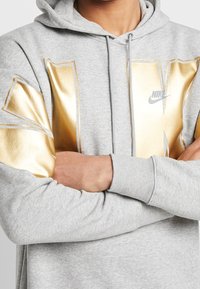 Grå hoodie med guld grafisk design på bröstet och ärmarna. Har dragsko i huvan och ribbade muddarna. Nike-logotyp på framsidan.