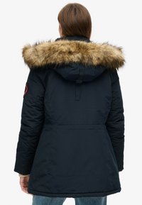 Navy insulated parka with a detachable faux fur hood, elastic cuffs, and a waist cinch for shape, featuring a patch on the left sleeve.
