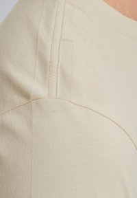 Beige bomulds t-shirt med rund halsudskæring og korte ærmer; synlig syning ved skulderen og sidesømmen.
