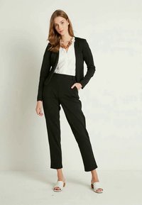 Mulher vestindo um blazer e calças pretas com uma blusa branca, colar robusto castanho e sandálias de salto abertas brancas, em pé contra um fundo simples.