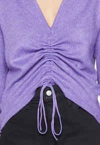 Fioletowy sweter dziany z dekoltem w serek i marszczeniami w talii, wyposażony w sznurek do ściągania. W połączeniu z czarnymi spodniami i guzikiem.