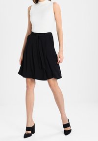 Jupe noire en forme de A avec un détail de taille nouée, associée à un haut sans manches blanc à côtes et des chaussures à talons noirs. Texture de tissu lisse.