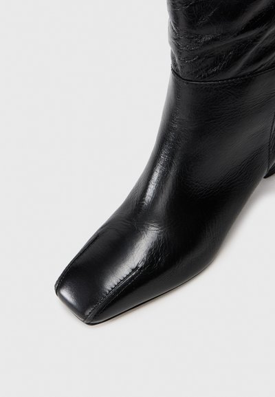 Fekete bőr, középig érő csizma, négyzetes orral és kissé ráncolt felső résszel, sima világos háttéren.