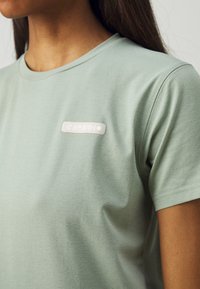 Tricou din bumbac de culoare verde deschis, cu guler rotund și mâneci scurte. Prezintă o mică etichetă albă ridicată cu logo pe pieptul stâng.