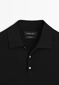 Svart bomullspikétröja med tre vita knappar och uppvikt krage, med en "Massimo Dutti"-märkesetikett inuti halsringningen.