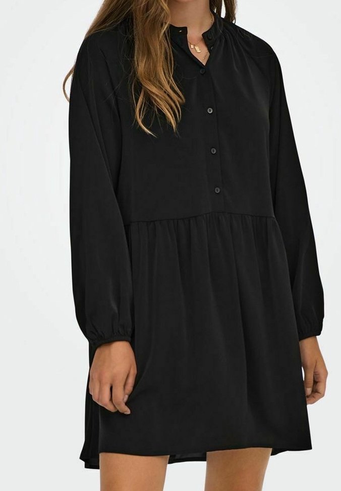 Femme portant une robe noire à manches longues avec des boutons sur le devant et une taille froncée, debout devant un fond uni.
