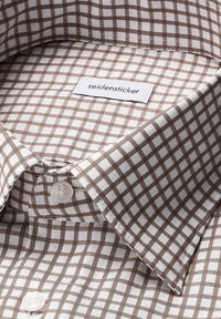 Colletto di camicia a quadri marroni e bianchi con etichetta. Presenta un bottone e una texture di cotone liscia, evidenziando un preciso motivo a griglia.