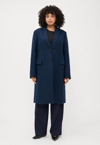 Donna dai capelli ricci in un cappotto blu navy sta in piedi con sicurezza, indossando una maglia nera e pantaloni a gamba larga, su uno sfondo bianco, semplice.