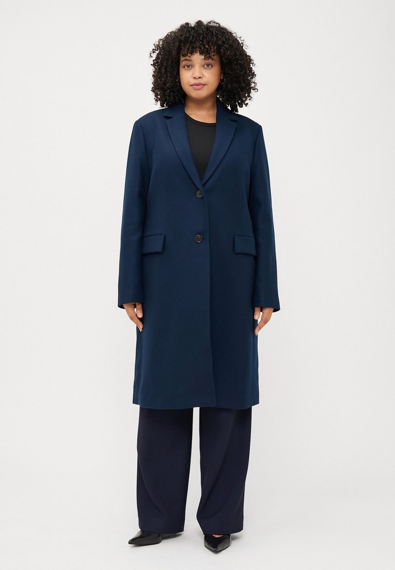 Donna dai capelli ricci in un cappotto blu navy sta in piedi con sicurezza, indossando una maglia nera e pantaloni a gamba larga, su uno sfondo bianco, semplice.