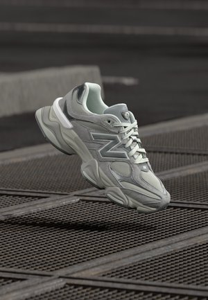 Γκρι αθλητικό παπούτσι New Balance με λευκά κορδόνια, πλέγμα και πάνελ από σουέντ, να αιωρείται πάνω από μια μεταλλική σχάρα σε εξωτερικό χώρο.