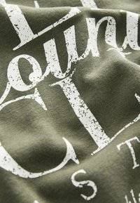 Alyvuogių žalia medvilnės audinys su baltu nusidėvėjusiu užrašu. Artimas planas rodo minkštą tekstūrą ir išlenktus kraštus, pabrėžiančius spausdinimo detales.