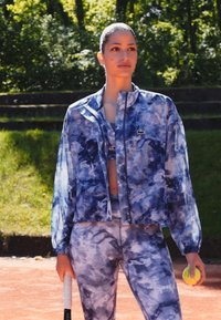 Modrá tie-dye windbreaker přes odpovídající crop top a legíny, se znakem Lacoste. Modelka drží tenisovou raketu a míček.