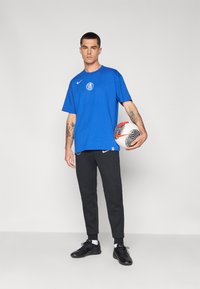 Blå Nike t-shirt med rundt logo, sorte Nike joggers og sorte træningssko. Holder en hvid og rød mønstret fodbold.