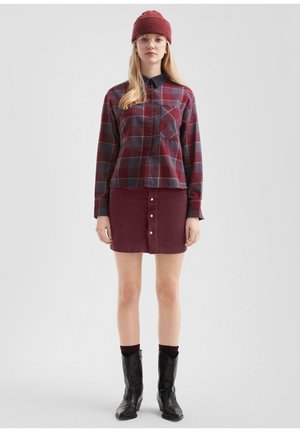Jeune femme portant une chemise à carreaux rouges, une jupe bordeaux avec des boutons, un bonnet assorti, des lunettes, des chaussettes foncées et des bottes noires, debout sur un fond uni.