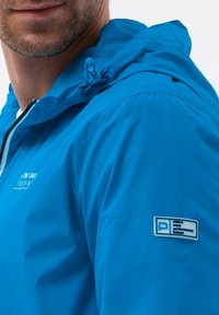 Blå letvægtsjakke med hætte, med logo-mærke på ærmet og en åndbar mesh-inderside. Glat, vandafvisende stof.
