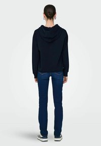 Felpa nera con cappuccio e maniche lunghe, abbinata a jeans skinny blu scuro. Il tessuto sembra morbido, con una vestibilità casual e dettagli minimi.